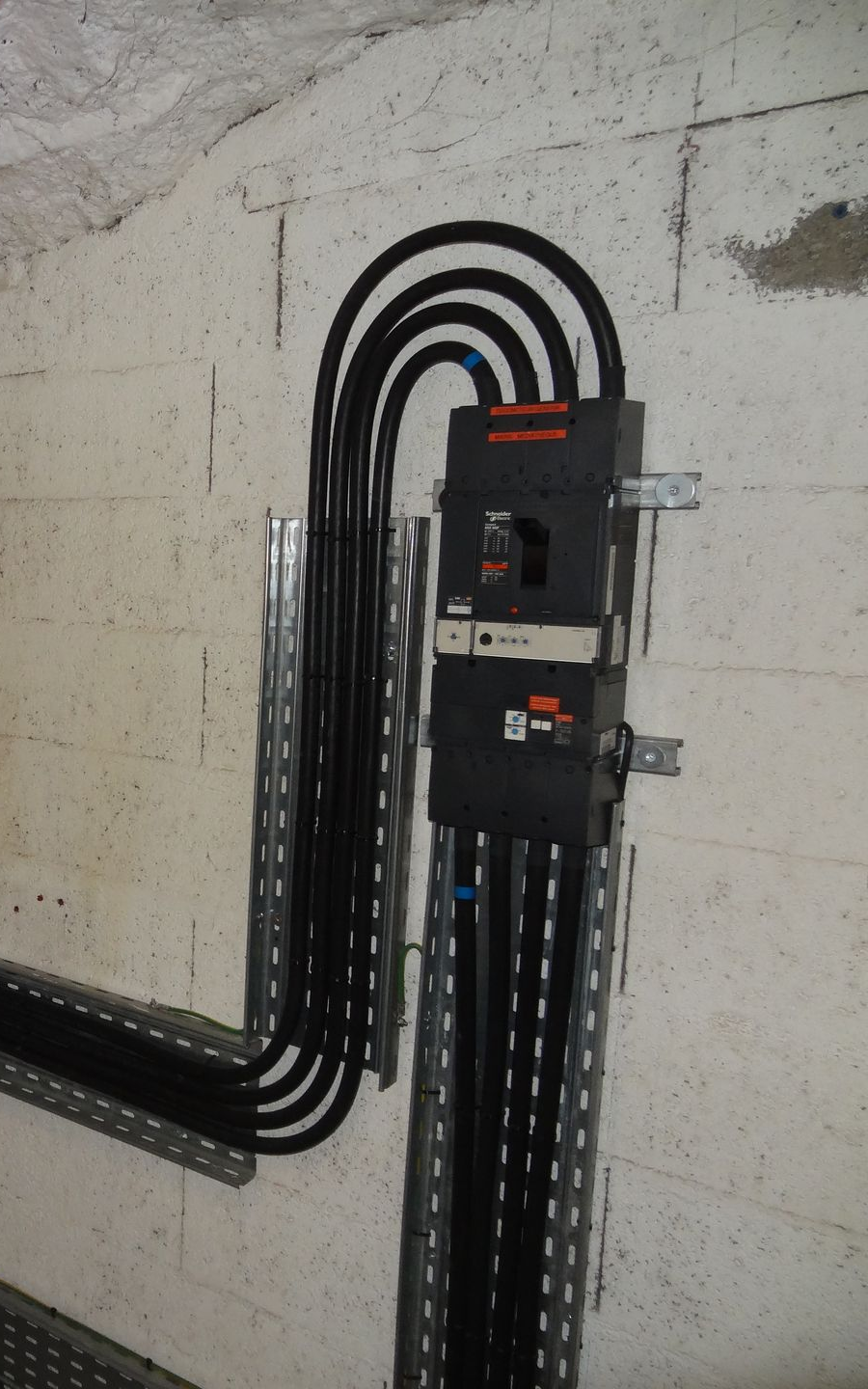 Disjoncteurs et câblage électrique fixés sur un mur en béton. Des câbles noirs passent dans des chemins de câbles métalliques.