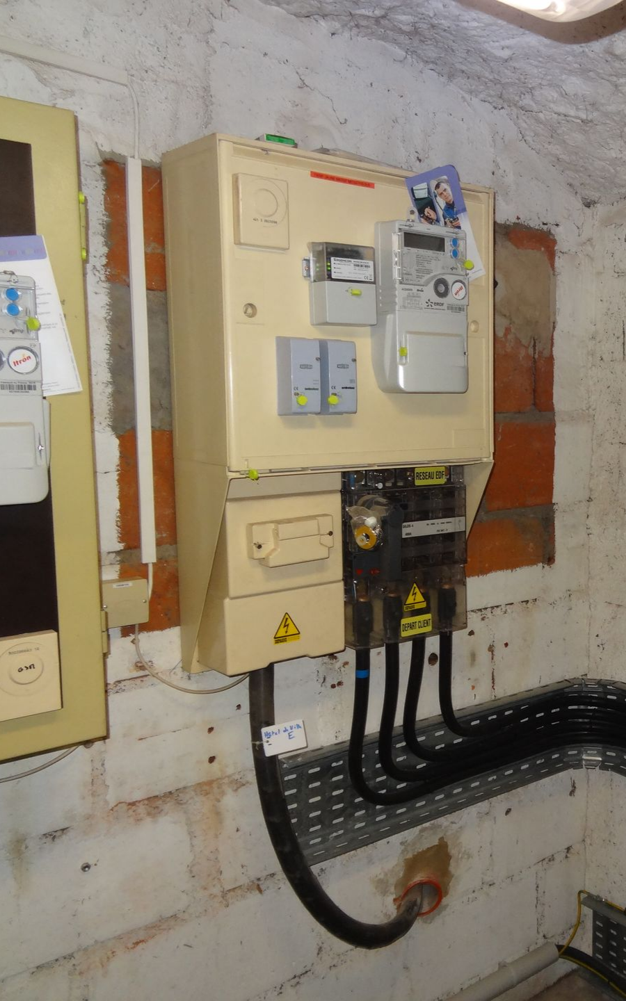 Boîtier de compteur électrique sur un mur de briques, avec des fils et un symbole d'avertissement.