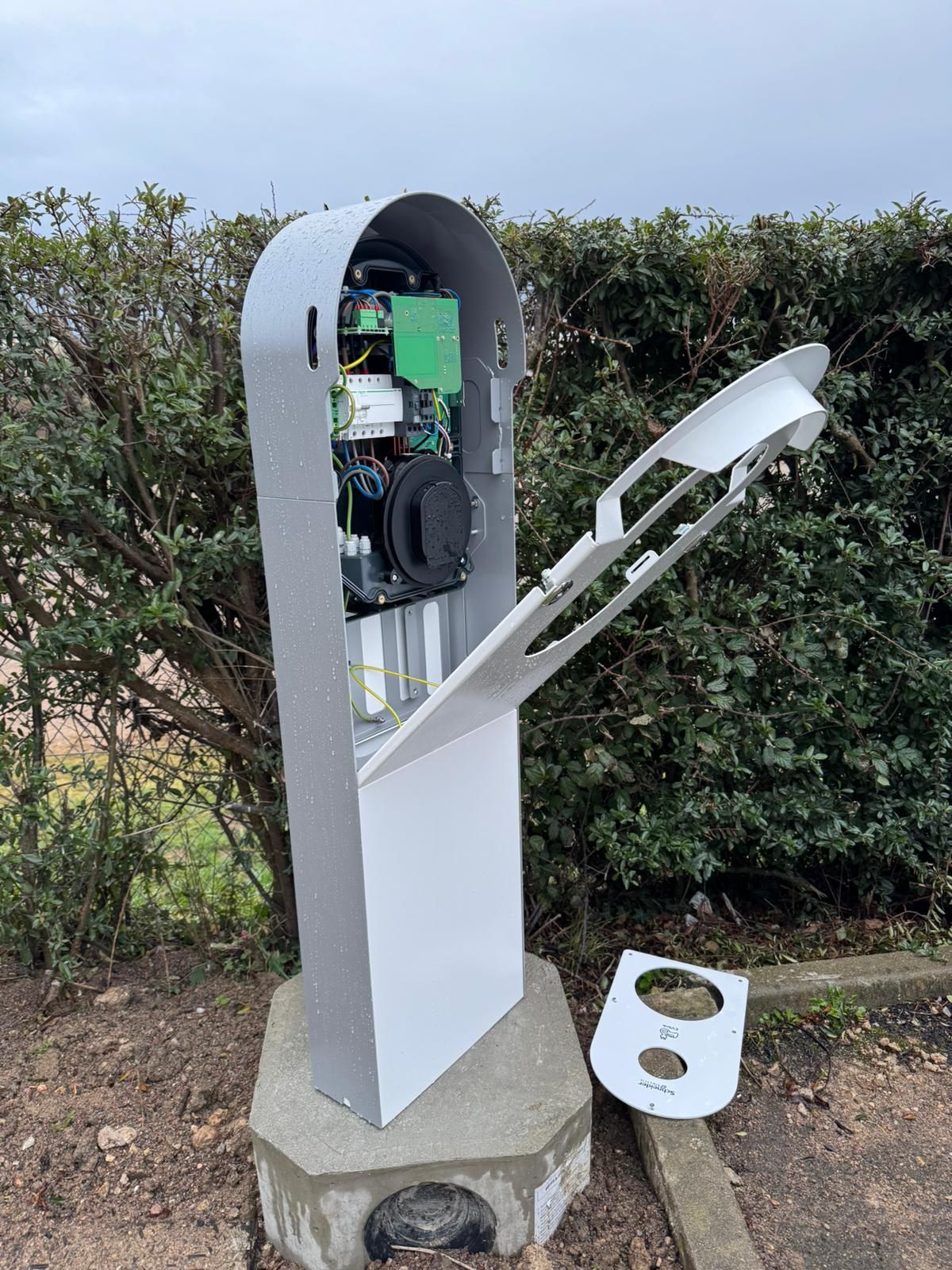 Voiture électrique blanche en charge dans une allée, à côté d'une maison entourée d'arbres et baignée de soleil.