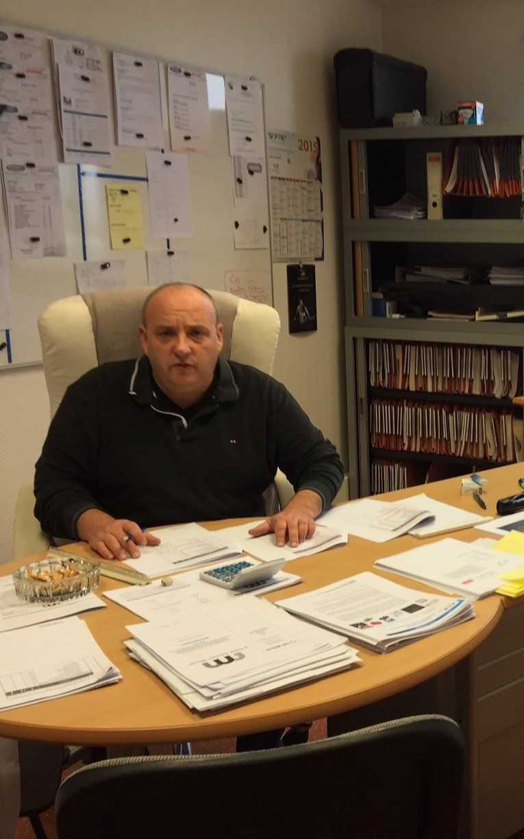 Un homme est assis à son bureau, regardant l'objectif. On aperçoit des papiers, un cendrier et des documents. En arrière-plan, un mur couvert de paperasse et une étagère.
