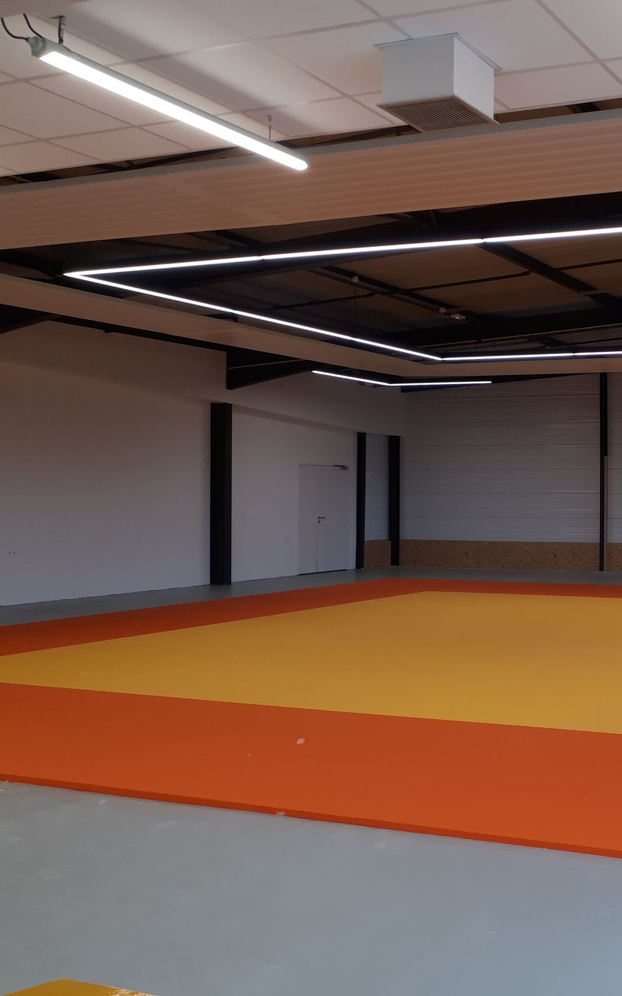 Espace vide pour la pratique de la lutte ou des arts martiaux, avec tapis orange et jaune et éclairage zénithal.
