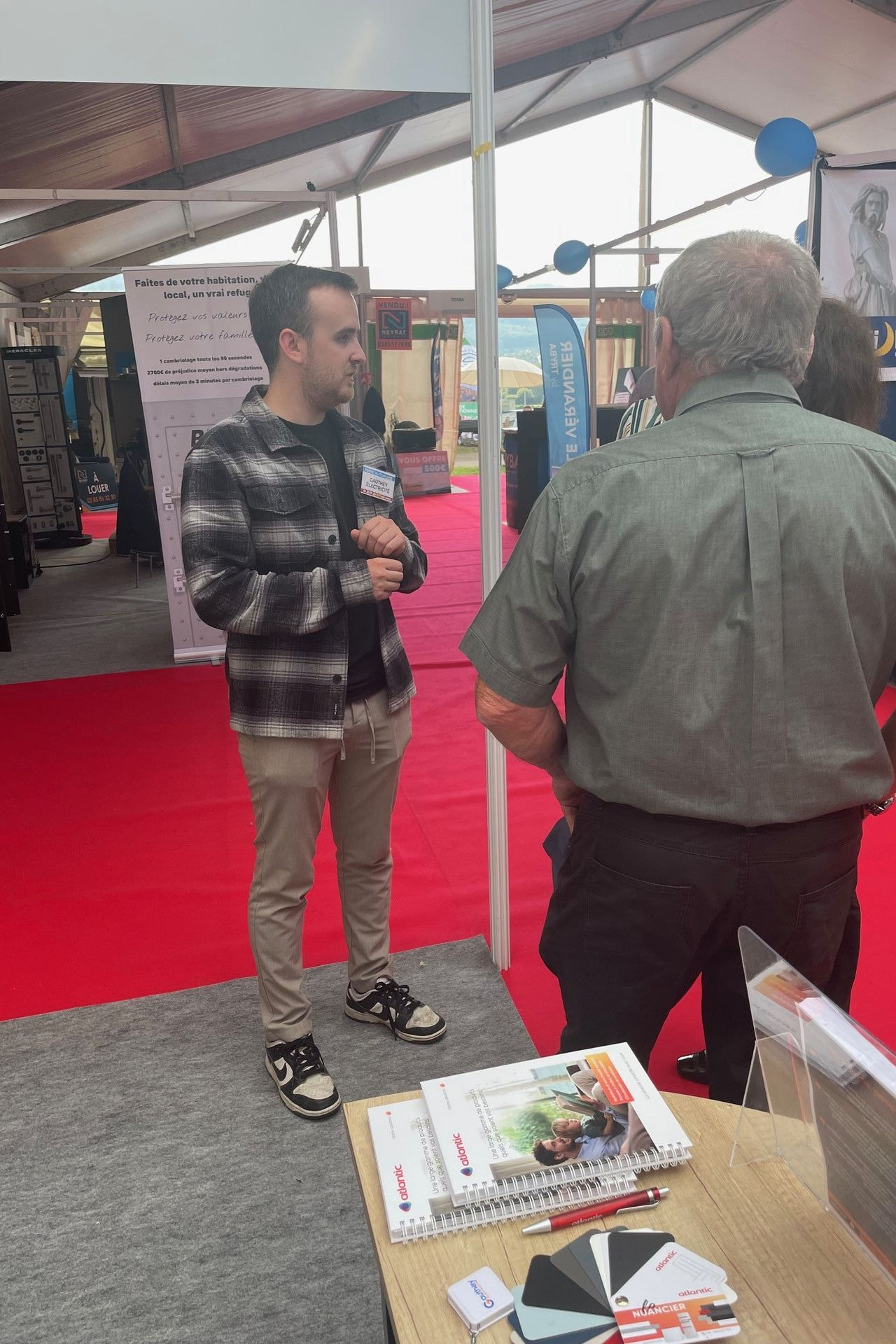 Un homme en chemise à carreaux discute avec un autre homme dans une cabine avec tapis rouge.
