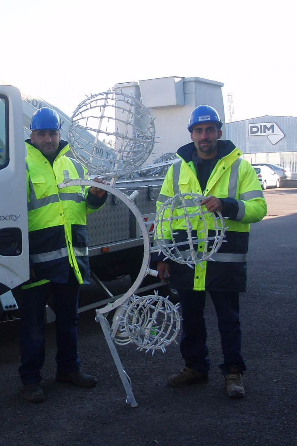 Deux hommes portant des gilets de sécurité et des casques de chantier tiennent des décorations sphériques en métal près d'un camion.