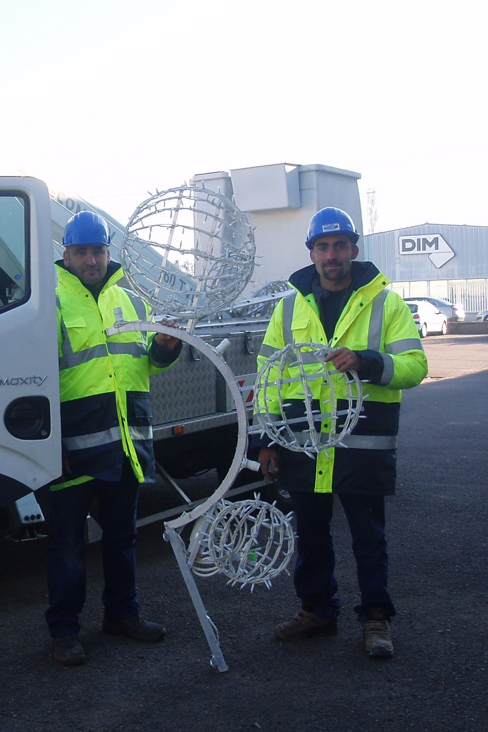 Deux hommes portant des gilets haute visibilité tiennent des structures lumineuses décoratives à l'extérieur, près d'un camion et d'un bâtiment sur lequel est inscrit « DIM ».