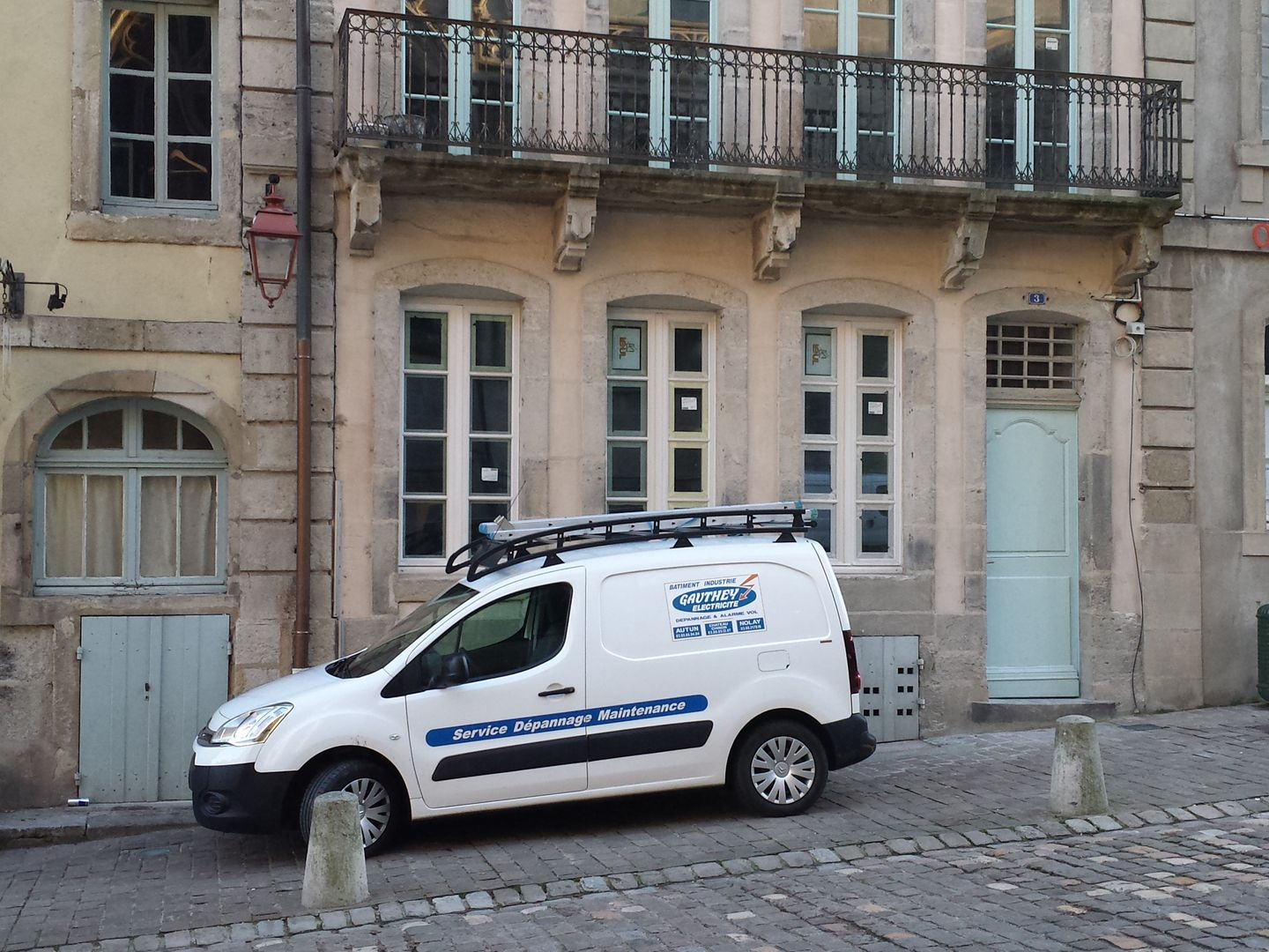 Une camionnette de service blanche est garée dans une rue pavée, devant un bâtiment de couleur claire.