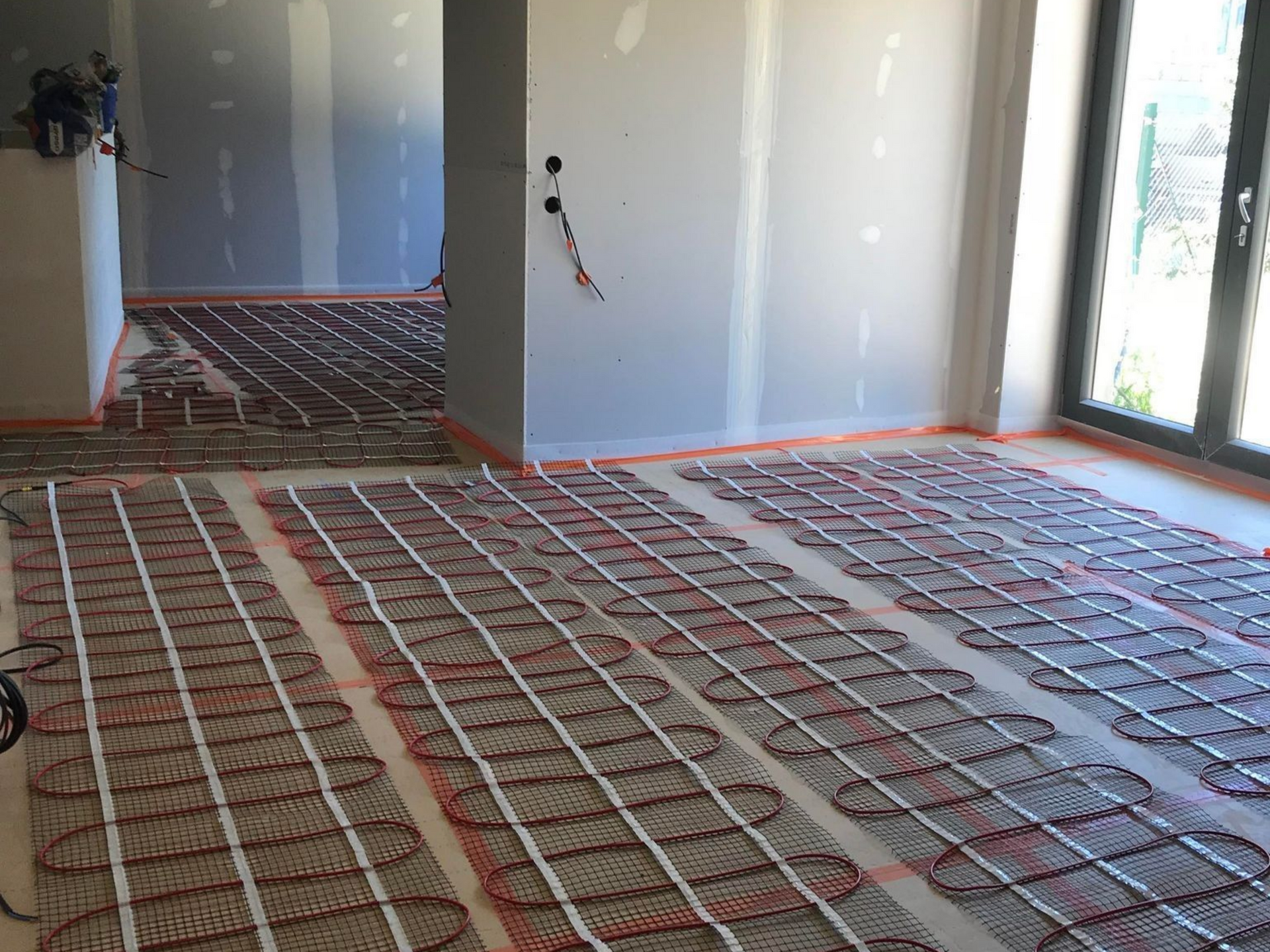 Pièce en travaux avec murs blancs, tuyaux apparents au sol et prises électriques.