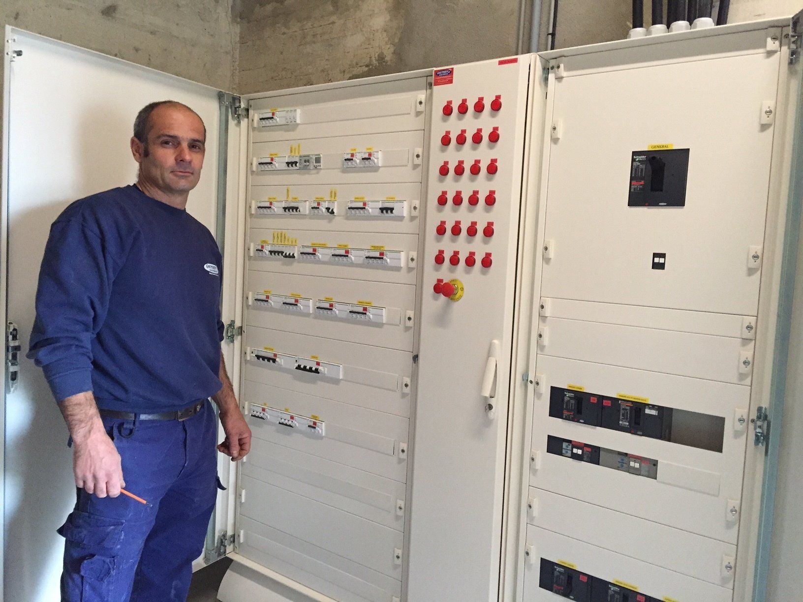 Un homme se tient à côté d'un tableau électrique ouvert comportant des rangées d'interrupteurs et de disjoncteurs ; scène en intérieur.