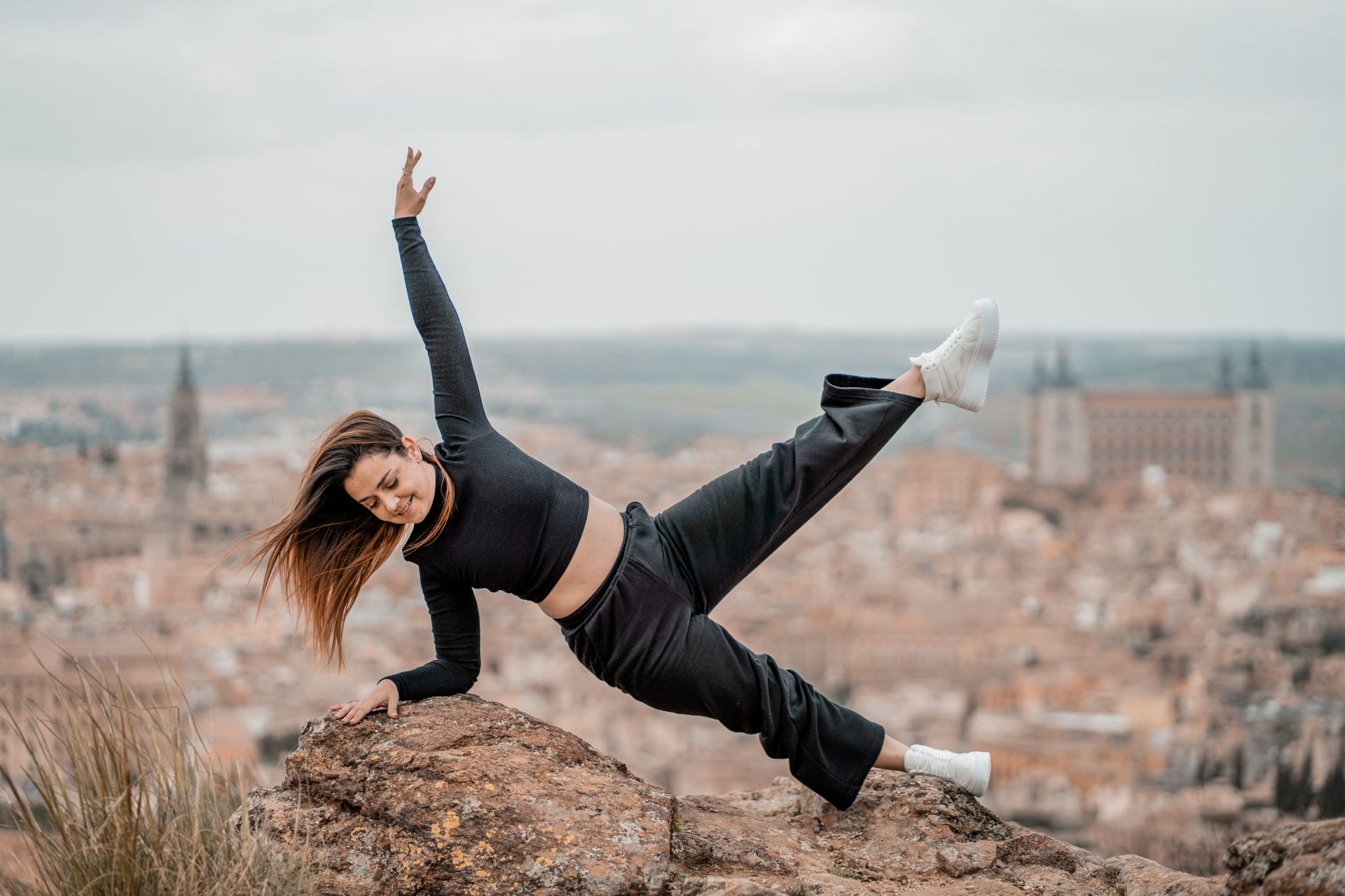 Mujer haciendo la plancha lateral sobre una roca, con el brazo levantado. Vestida de negro y con zapatos blancos, con vistas