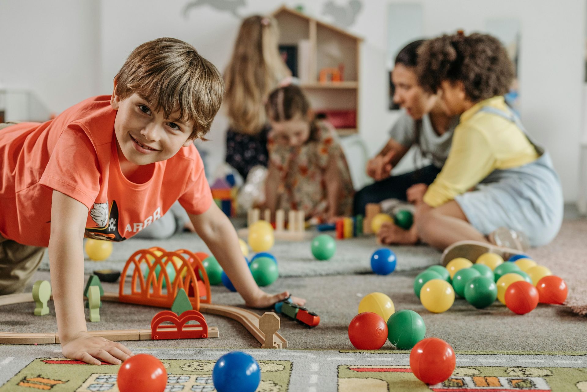 Niño sonriente, jugando con un tren de juguete entre pelotas de colores, en una sala de juegos con otros niños y un adulto.