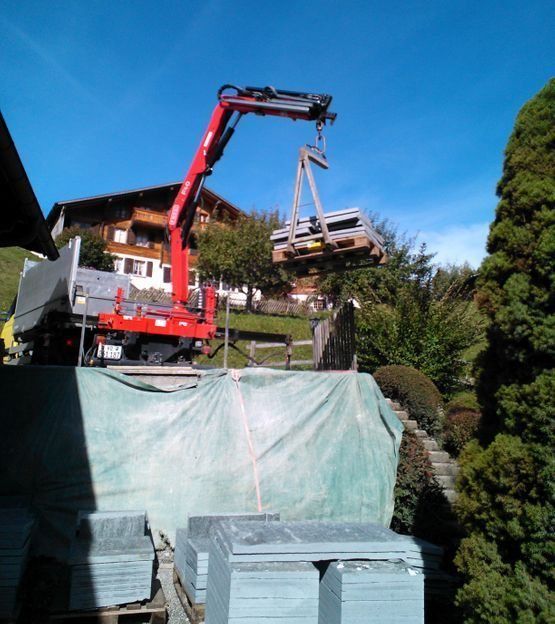 Une grue rouge soulève une palette avec une maison en arrière-plan