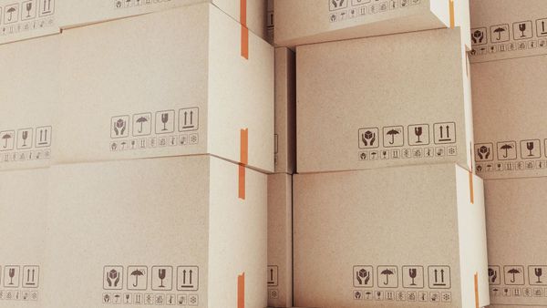 Cajas de cartón beige apiladas con cinta naranja, dentro de un almacén.