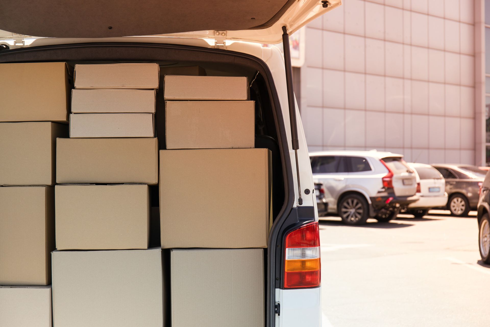 Furgoneta blanca abierta por detrás, llena de cajas de cartón apiladas, estacionada afuera con autos.