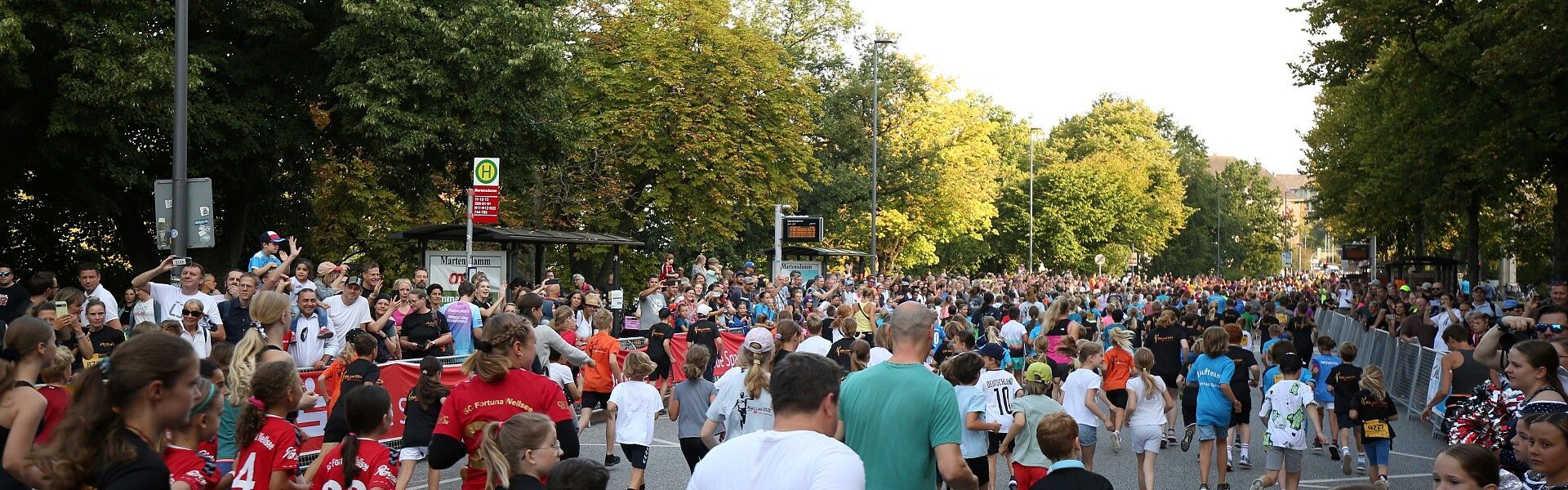 Eine Menschenmenge aus Läufern und Zuschauern versammelt sich auf einer von Bäumen gesäumten Straße an der Startlinie eines Straßenlaufs.