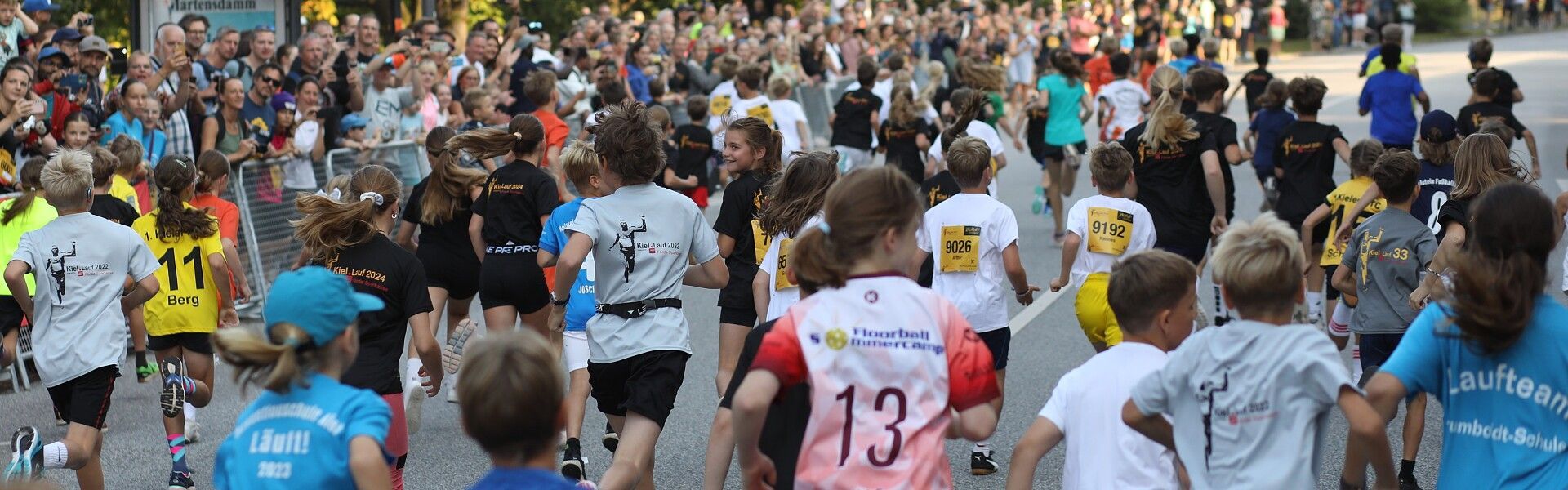Läufer und Zuschauer drängen sich bei einem Straßenlauf; von hinten sind die Teilnehmer in ihren farbenfrohen T-Shirts zu sehen.