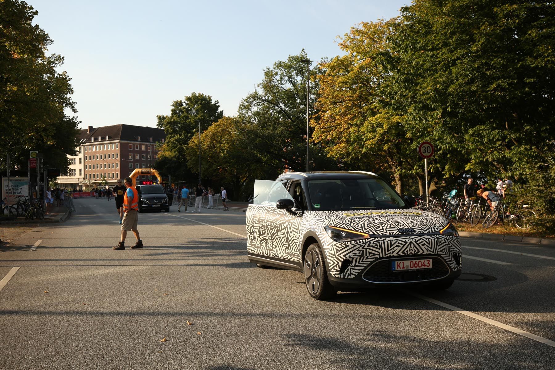 Ein getarnter Geländewagen fährt auf einer von Bäumen gesäumten Straße, in deren Nähe eine Person die Straße überquert.