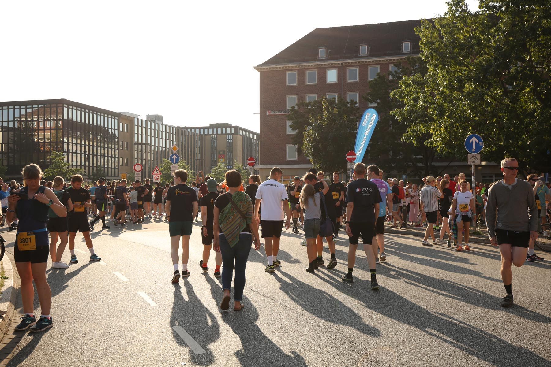 Läufer und Spaziergänger versammelten sich auf einer sonnigen Straße an der Startlinie eines Rennens, in der Nähe von langen Schatten und Backsteingebäuden.