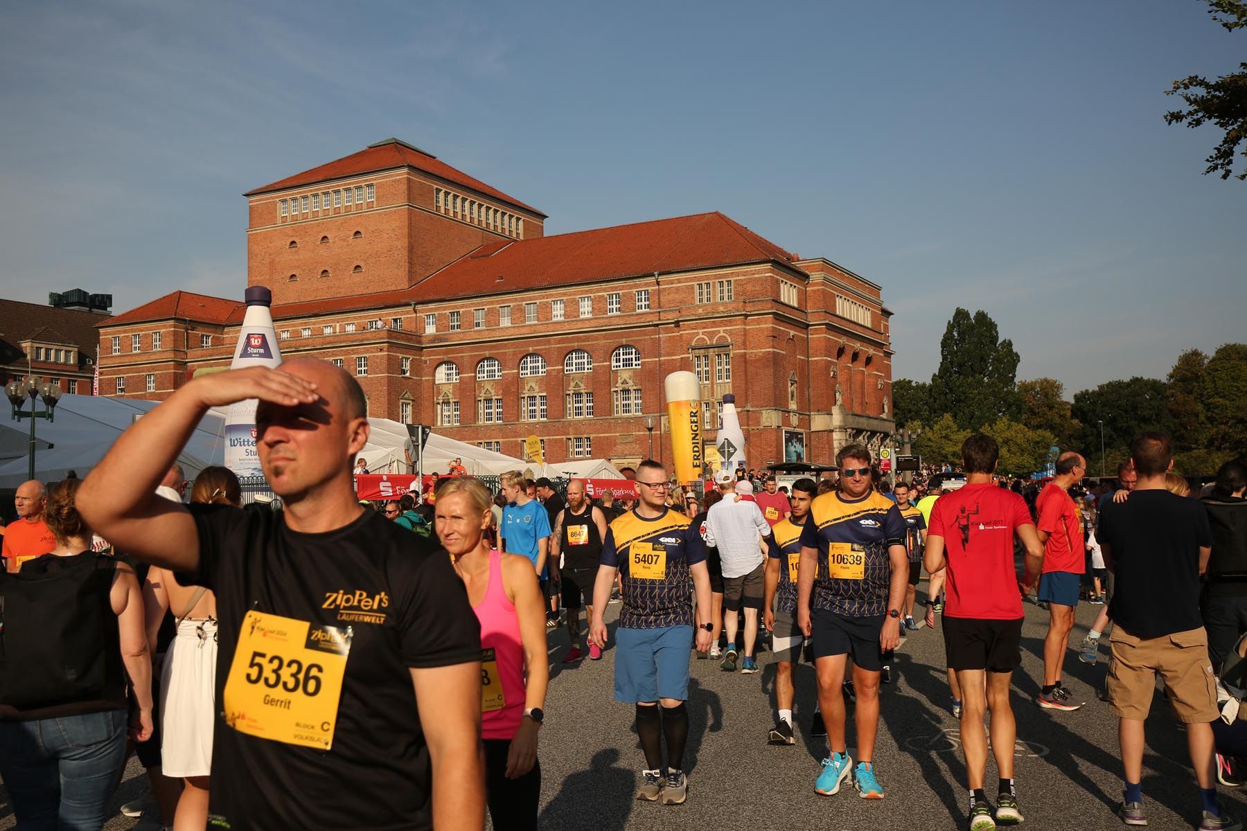 An einem sonnigen Tag versammeln sich Läufer an einem Startbereich vor einem Backsteingebäude.