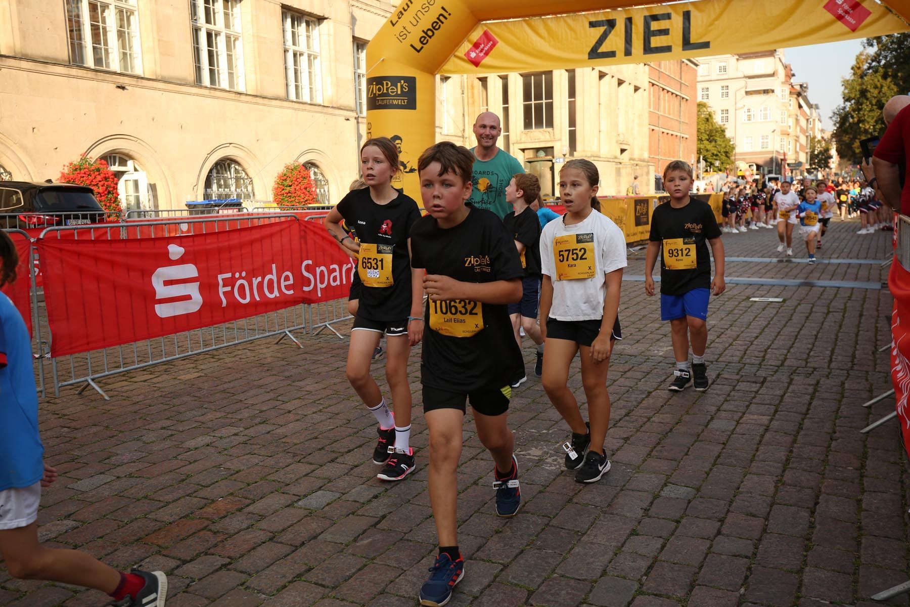 Läufer in schwarzen T-Shirts nähern sich der Ziellinie unter einem gelben „ZIEL“-Banner auf einer Kopfsteinpflasterstraße.