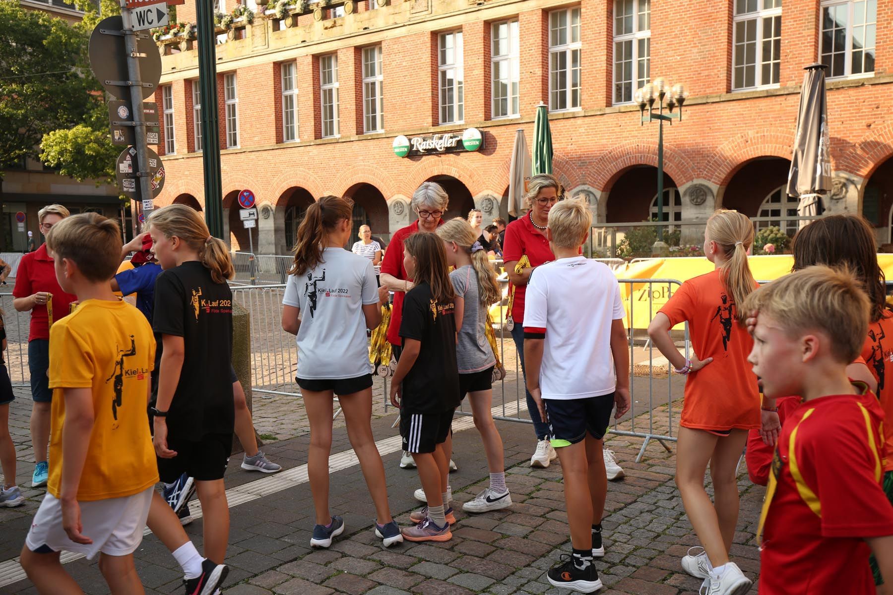 Eine Gruppe von Menschen in farbenfroher Sportkleidung geht auf einer Kopfsteinpflasterstraße neben Backsteingebäuden entlang.