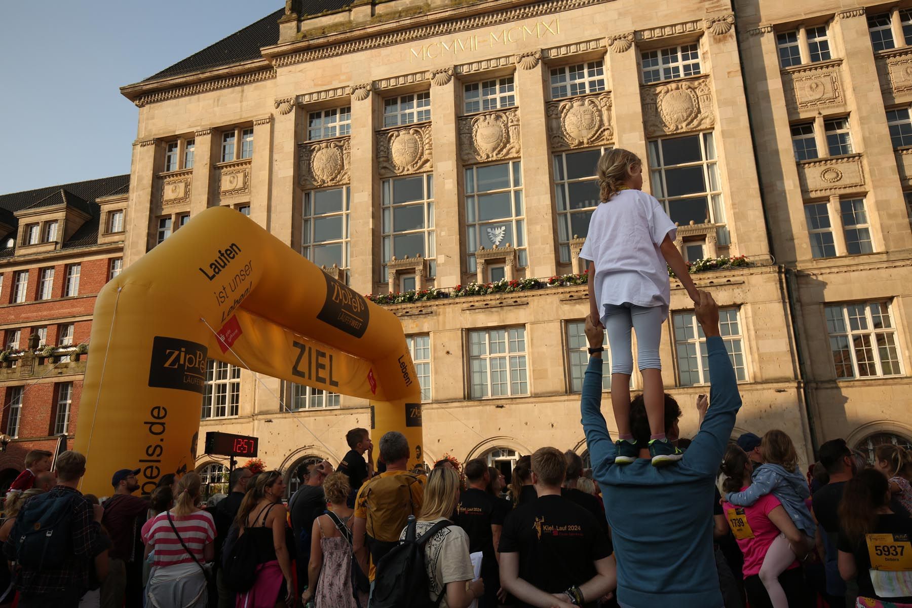 Menschenmenge vor einem Steingebäude mit einem gelben aufblasbaren Bogen und einem Kind auf den Schultern von jemandem.