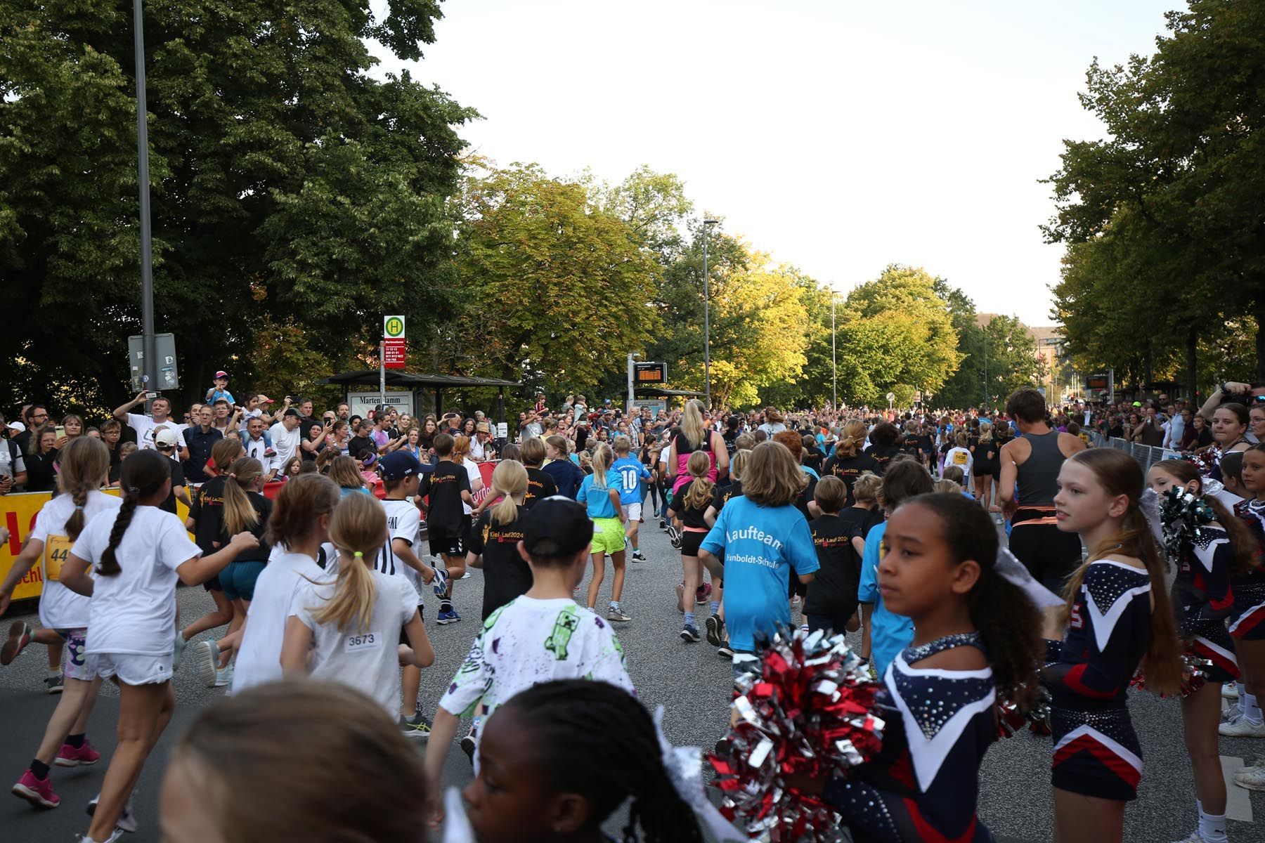 Auf einer von Bäumen gesäumten Straße versammelte sich eine Menschenmenge zu einer Parade mit Tänzern und Cheerleadern in weißen und marineblauen Uniformen.