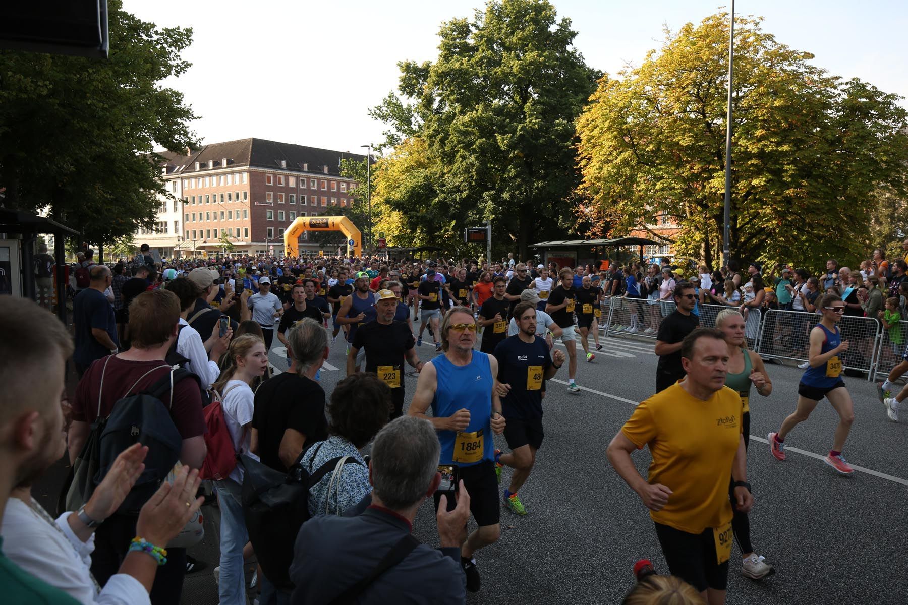 Ein gut besuchtes Freiluftrennen, bei dem die Läufer in der Abenddämmerung an den Zuschauern auf einer von Bäumen gesäumten Straße vorbeiziehen.
