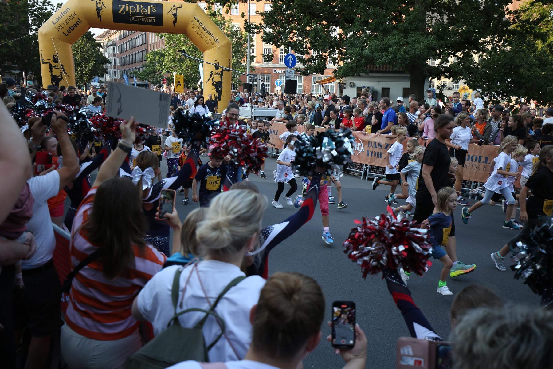 Cheerleader treten bei einer Straßenparade auf, die von einer Zuschauermenge unter einem gelben Bogen verfolgt wird.