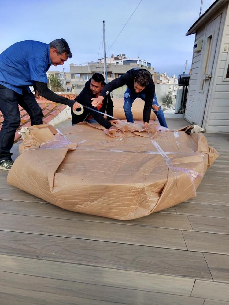 Tres personas abren un gran paquete de cartón sellado con cinta adhesiva en una terraza de madera junto al agua.