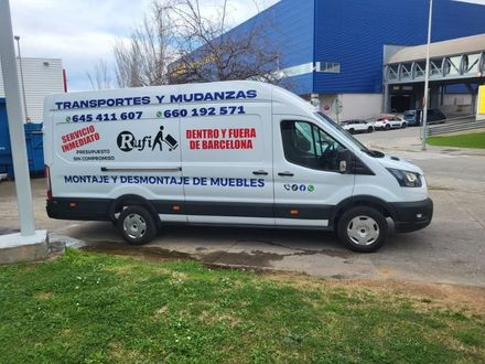 Furgoneta blanca de mudanzas con letreros de transporte español aparcada frente a un edificio y una valla azules.