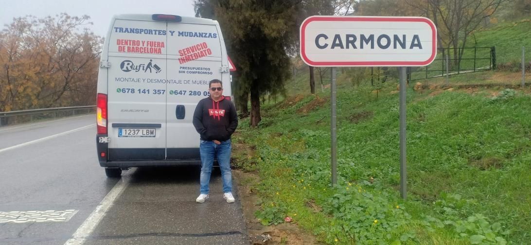 Persona de pie junto a una furgoneta blanca al lado de un cartel de carretera que dice “CARMONA”.