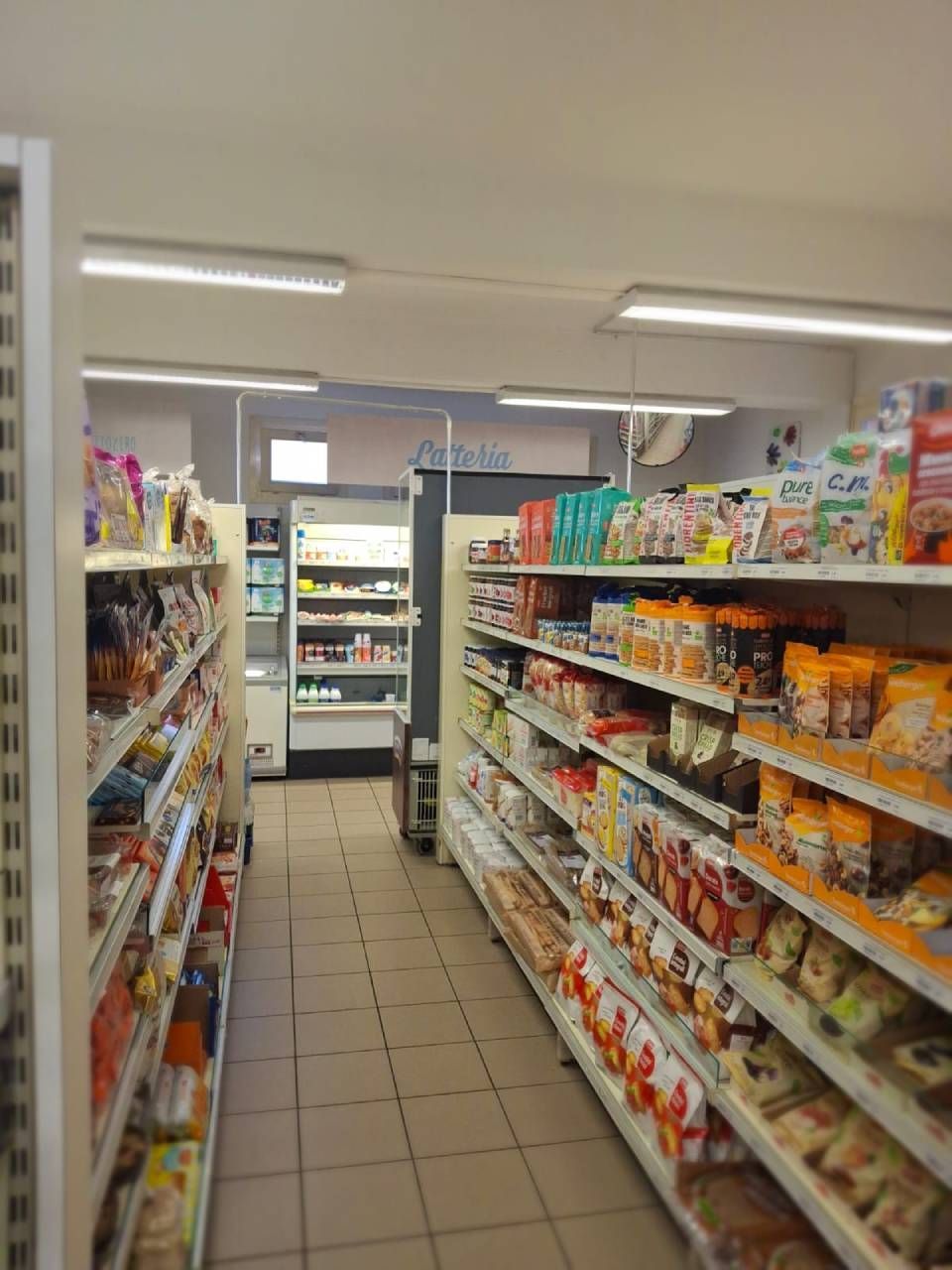 Interno di un minimarket, scaffali pieni di prodotti vari. Corsie con merce, luci bianche sul soffitto.
