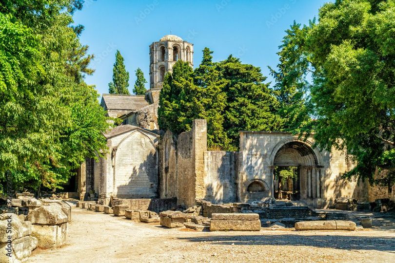 Des ruines de pierre antiques, avec des arches et une tour, se dressent au milieu d'arbres verdoyants, sous un ciel d'un bleu éclatant.