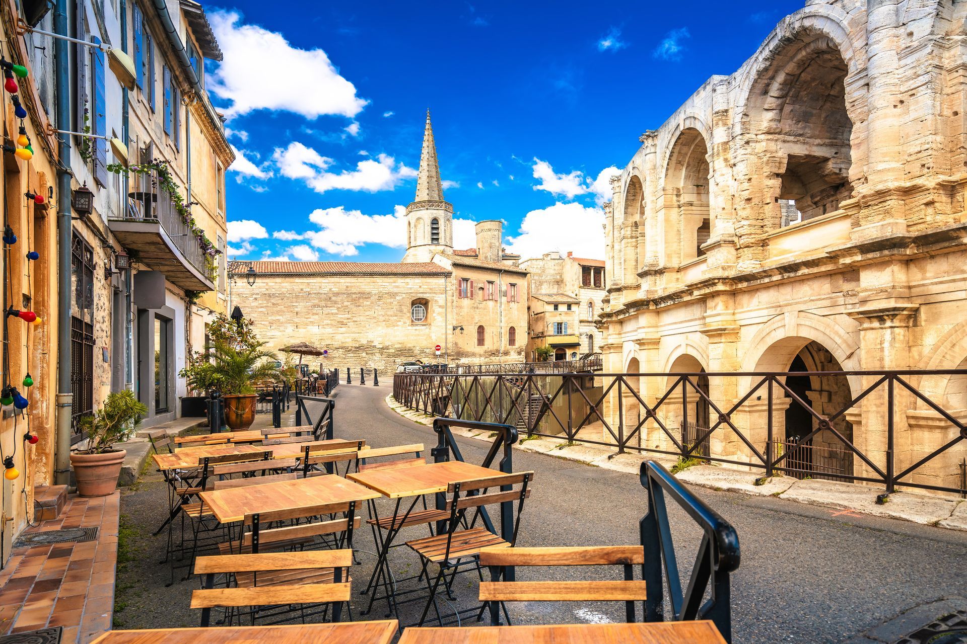 Des tables de café en terrasse jouxtent d'anciennes ruines de pierre sous un ciel d'un bleu éclatant, sur une place historique baignée de soleil.