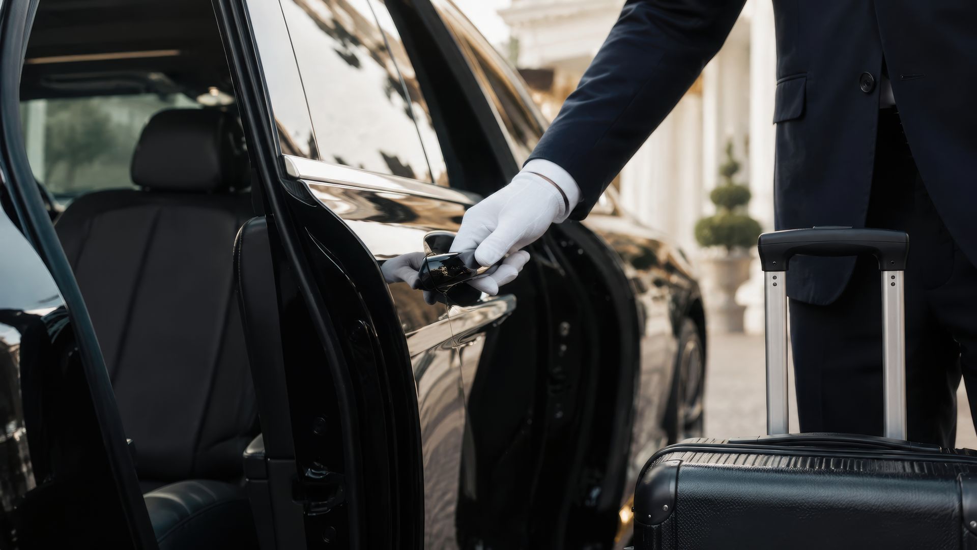 Un chauffeur ouvre la portière de sa voiture noire à côté d'une valise à l'entrée d'un hôtel.