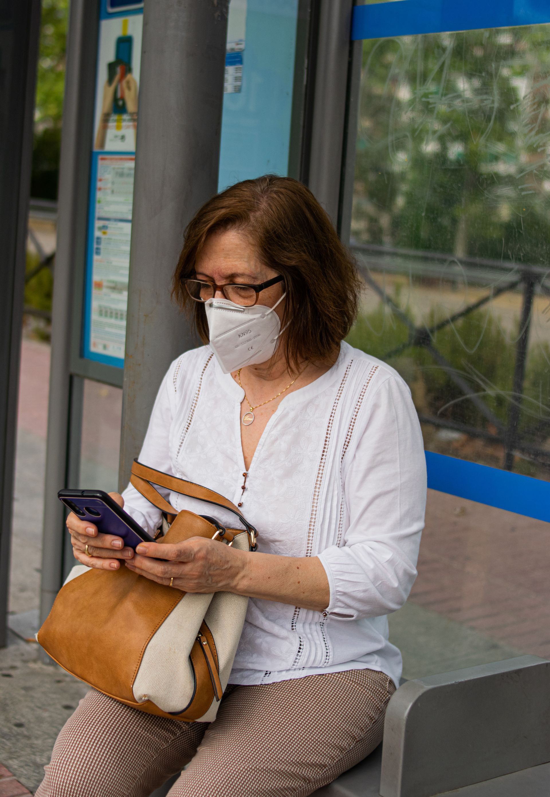 Une femme portant un masque est assise à un arrêt de bus, regardant son téléphone et tenant un sac beige.