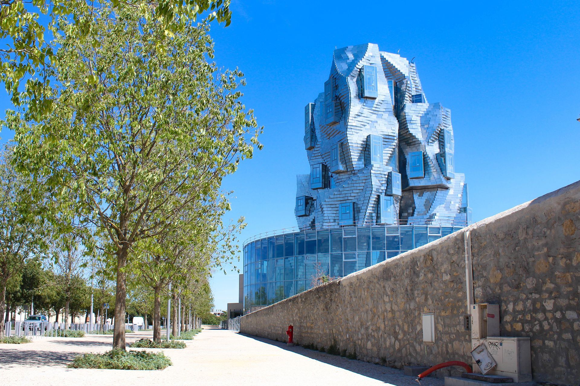 Bâtiment bleu miroir à côté d'un mur de pierre et d'une allée bordée d'arbres sous un ciel clair
