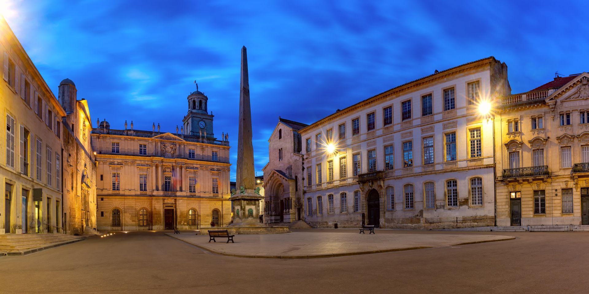 Vue du coucher de soleil sur une place historique européenne avec des bâtiments en pierre illuminés, une église et une haute flèche.