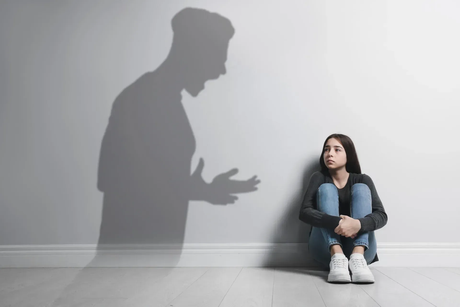 Una joven está sentada contra una pared, asustada, con la sombra de un hombre enojado sobre ella.