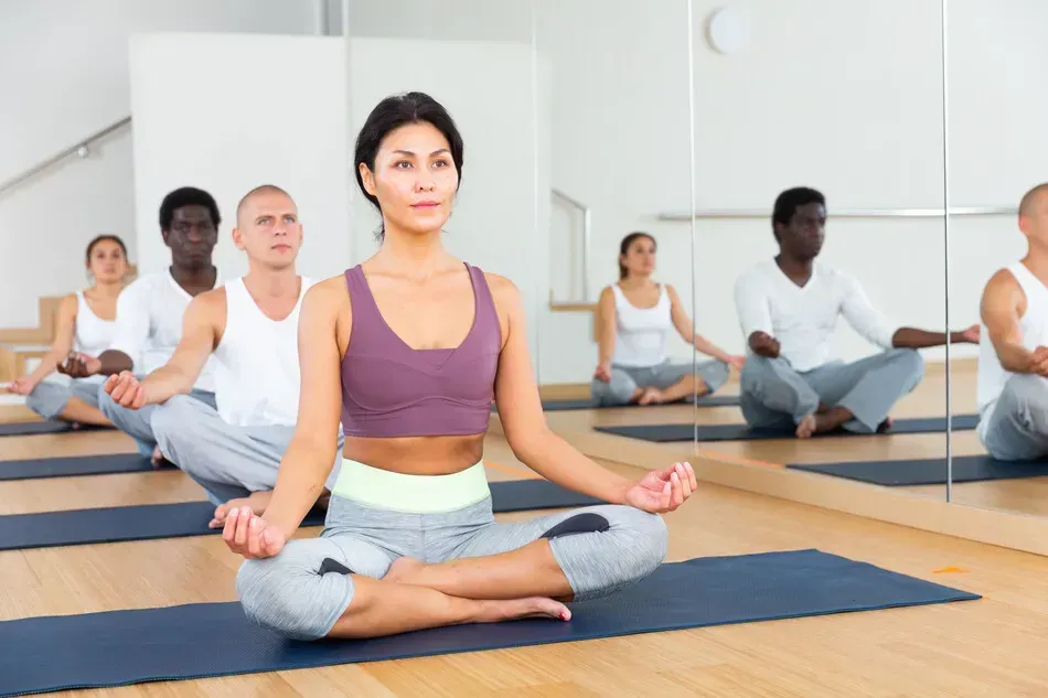 Grupo en clase de yoga, personas en posición de loto, expresiones neutrales, sala iluminada con espejos.