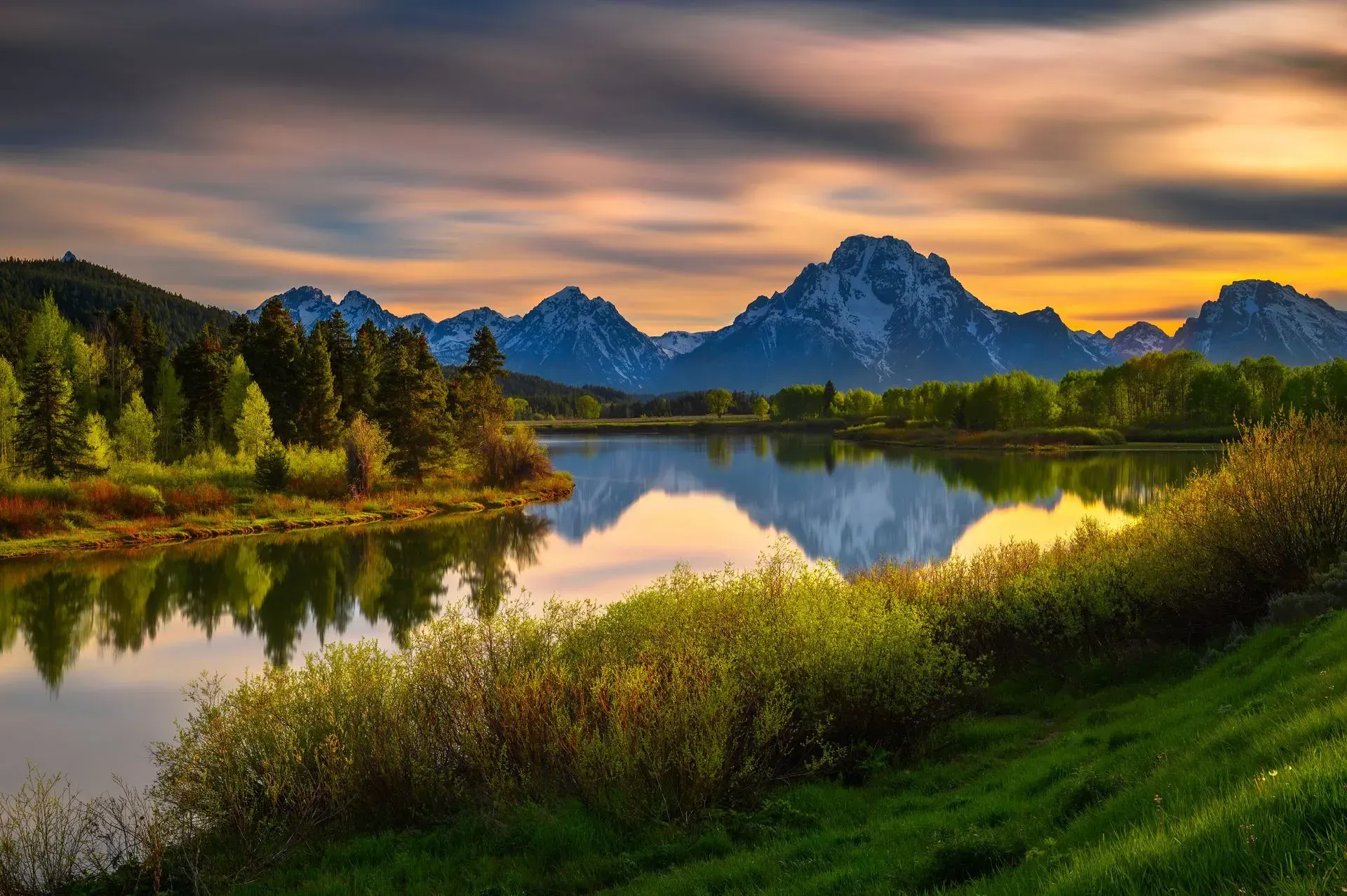 Lago que refleja las montañas al atardecer, con follaje verde y cielo dorado.