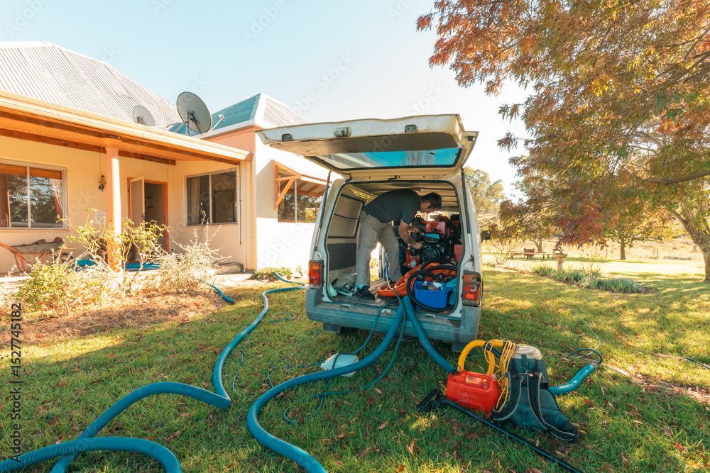 Ein Arbeiter saugt einen weissen Lastwagen in einem sonnigen Hof ab; auf dem Rasen liegen blaue Schläuche und Geräte.