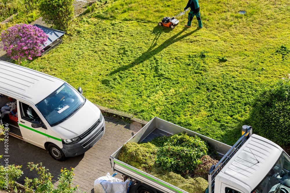 Zwei weisse Lieferwagen stehen vor einem grasbewachsenen Hof, in dem eine Person den Rasen mäht.