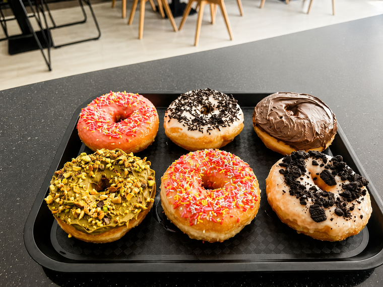 Seis donas variadas en una bandeja negra, cubiertas con chispas de colores, nueces, chocolate y glaseado.