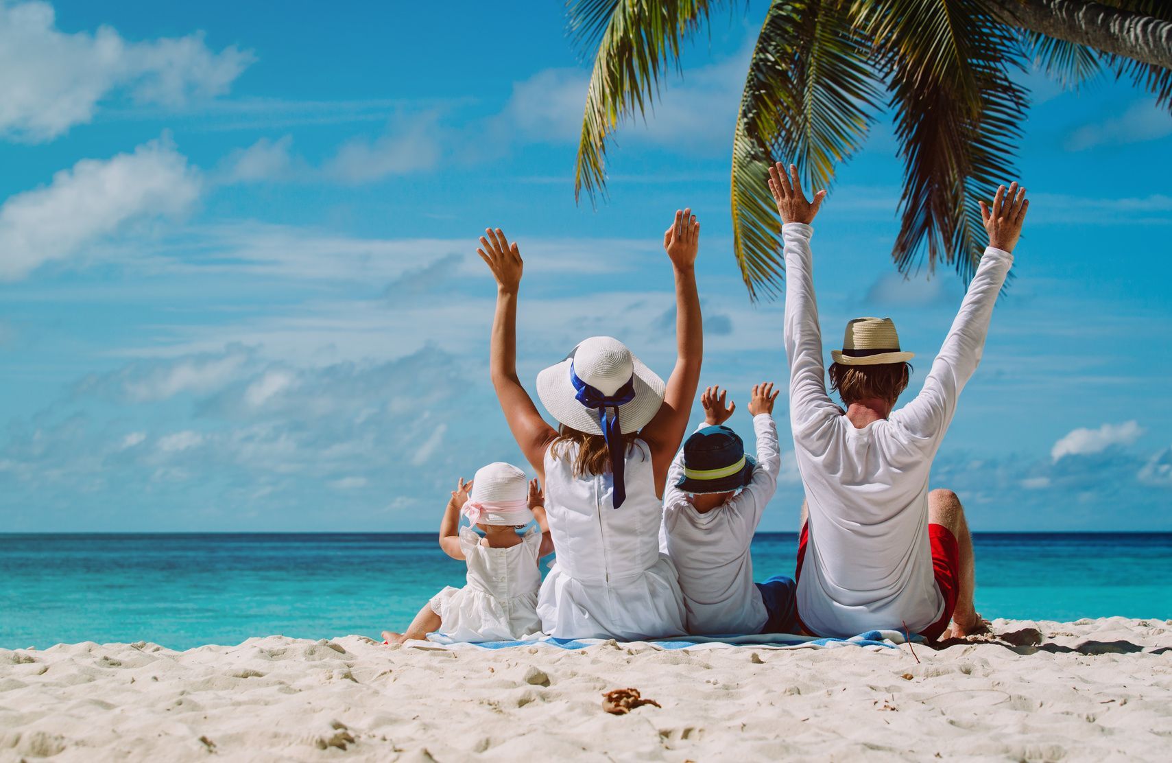 Une famille de quatre personnes est assise sur une plage de sable tropical, les bras levés, face à l'océan bleu, sous un palmier.