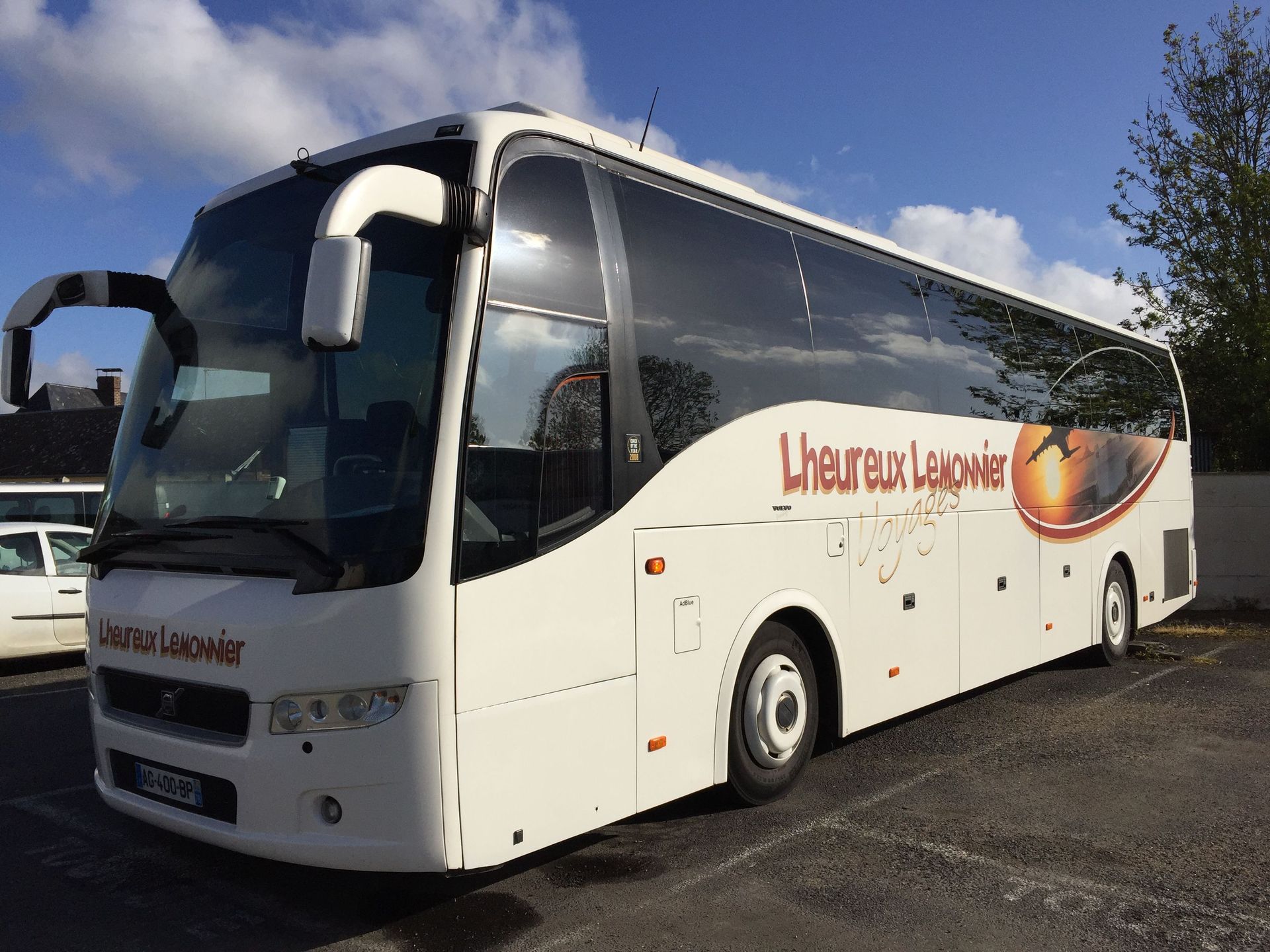 Un bus touristique blanc avec l'inscription « L'heureux Lehovier » sur le côté, garé sur un parking extérieur par une journée ensoleillée.