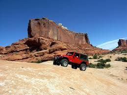 Red Jeep off-roading on a sandy trail in front of a large red rock formation under a blue sky.