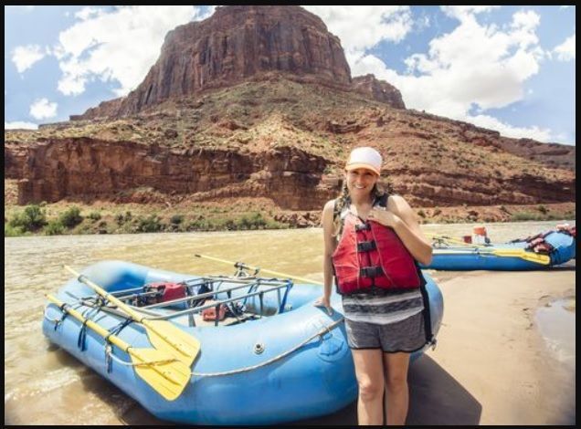 Woman in life vest stands near a blue raft on a riverbank with red rock cliffs in the background.