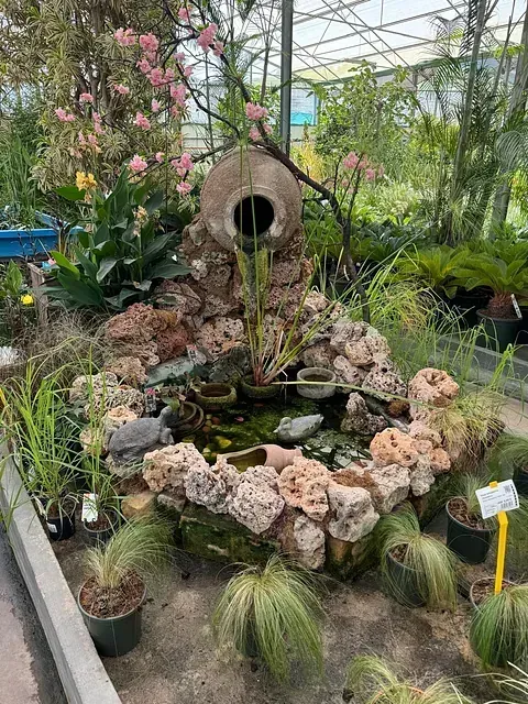 Una fuente de agua en un invernadero rodeada de plantas en macetas y flores.