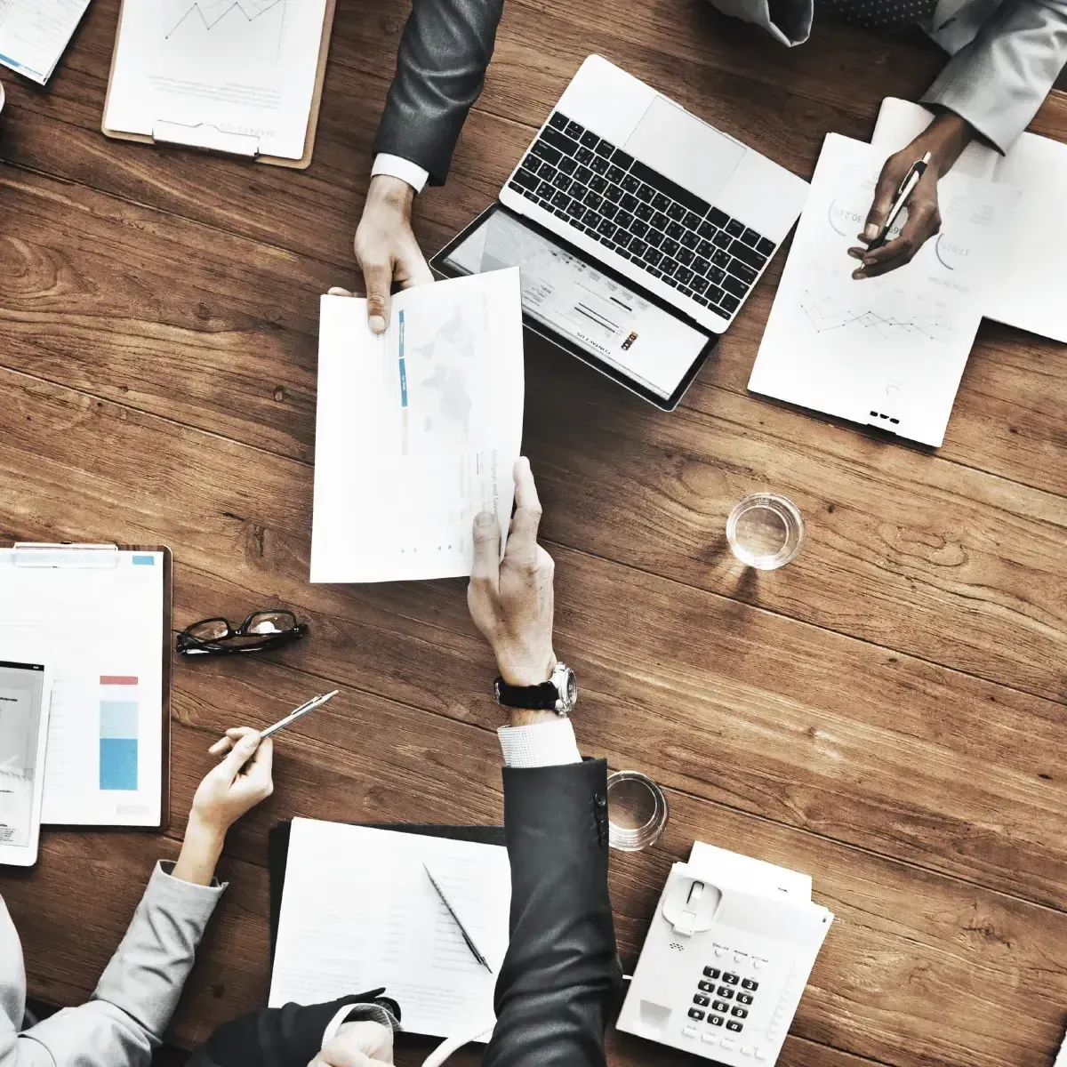Um grupo de pessoas está sentado ao redor de uma mesa de madeira com papéis e um laptop.