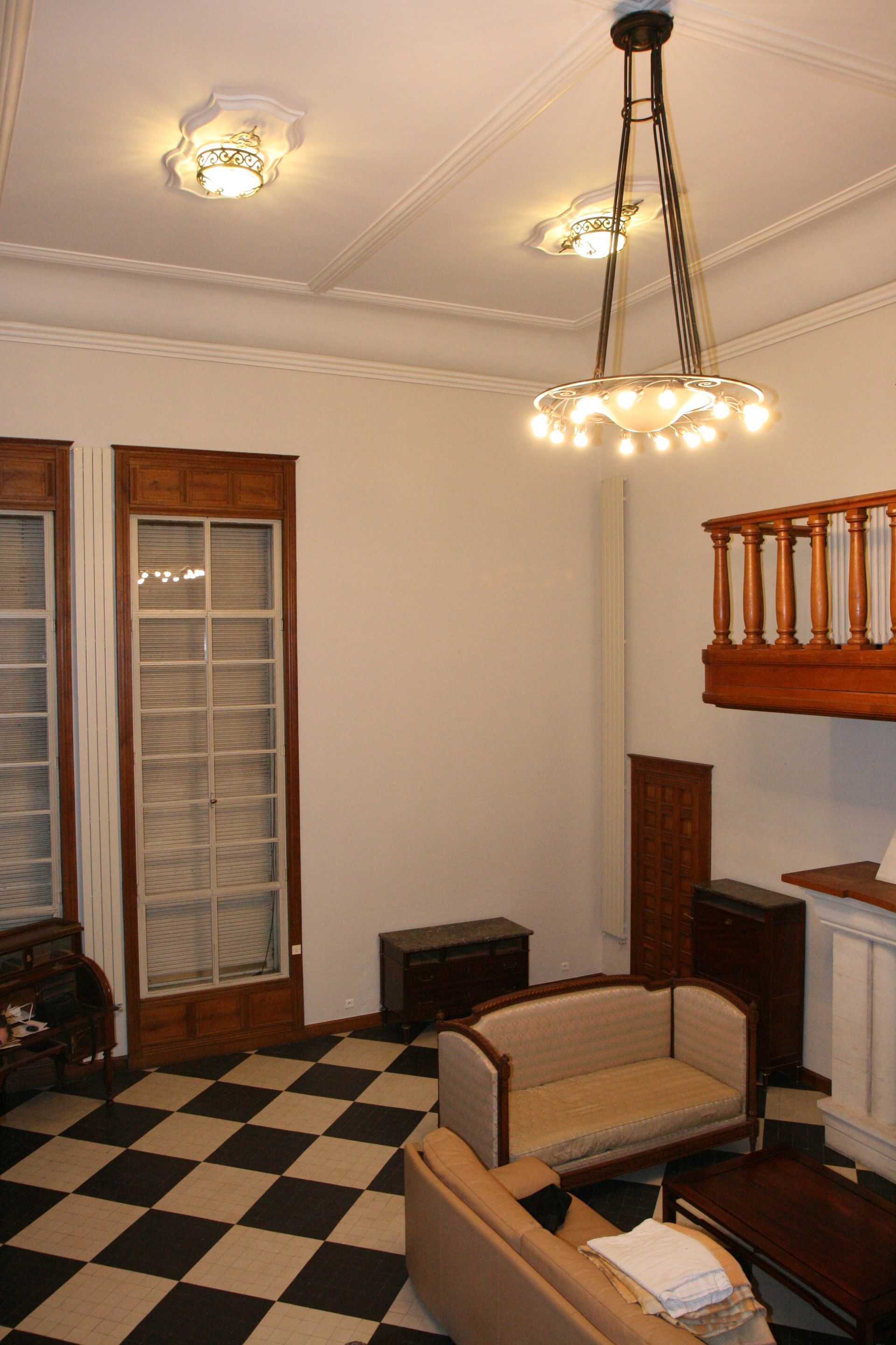 Chambre avec sol à damier, deux canapés, luminaires de plafond ornés, grandes fenêtres et petit balcon.